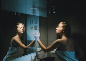A young woman wrapped in a towel touches the mirror in a contemporary bathroom setting.