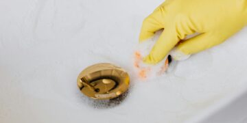From above of crop anonymous person in yellow rubber protective glove using sponge and detergent for washing white sink in bathroom