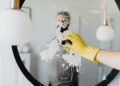 Anonymous male housekeeper in yellow rubber gloves wiping foam from round mirror in bathroom