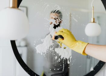 Anonymous male housekeeper in yellow rubber gloves wiping foam from round mirror in bathroom