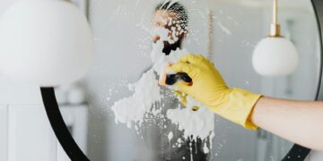 Anonymous male housekeeper in yellow rubber gloves wiping foam from round mirror in bathroom