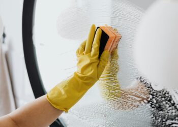 Anonymous person in yellow rubber gloves using simple sponge for cleaning mirror in bathroom