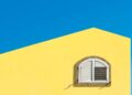 Bright yellow wall with an arched white shuttered window against a clear blue sky.