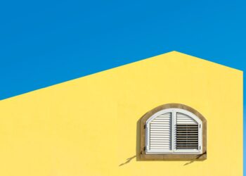 Bright yellow wall with an arched white shuttered window against a clear blue sky.