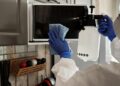 Individual in protective gear cleaning microwave using spray bottle and cloth indoors.