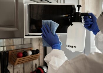 Individual in protective gear cleaning microwave using spray bottle and cloth indoors.