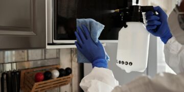 Individual in protective gear cleaning microwave using spray bottle and cloth indoors.