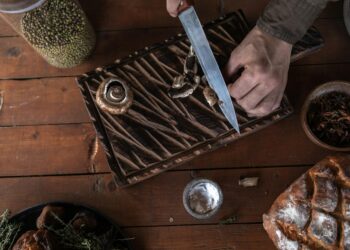 Top-down view of hands slicing mushrooms on a cutting board. Rustic wood setting.