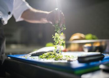Chef's hand preparing chopped green vegetables on a cutting board in a kitchen.