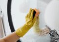 Anonymous person in yellow rubber gloves using simple sponge for cleaning mirror in bathroom