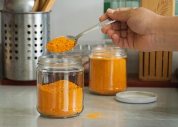 A close-up of turmeric powder being scooped into a jar in a kitchen with utensils.