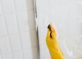 From above of crop anonymous person in yellow rubber protective glove washing shower cabin glass with sponge and detergent