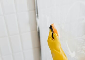 From above of crop anonymous person in yellow rubber protective glove washing shower cabin glass with sponge and detergent