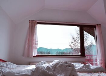 Bright bedroom interior with rumpled bed facing mountain view from window.