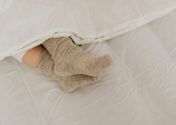 Comfortable feet in woolen socks peek out under a soft white linen blanket on a bed.