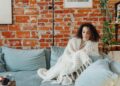 Woman wrapped in a white blanket relaxing on a sofa against a brick wall indoors.