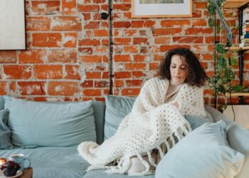 Woman wrapped in a white blanket relaxing on a sofa against a brick wall indoors.