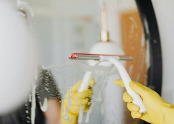 Anonymous person in yellow rubber gloves wiping water by squeegee from mirror in bathroom