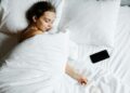 A young woman sleeps comfortably in bed with a smartphone beside her, wrapped in white sheets.