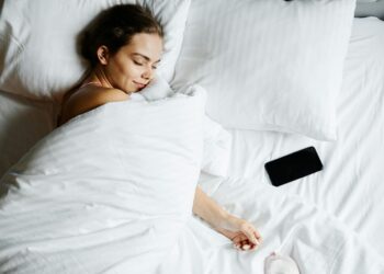 A young woman sleeps comfortably in bed with a smartphone beside her, wrapped in white sheets.
