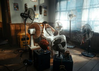 Man in white outfit sitting surrounded by several fans indoors during daytime.