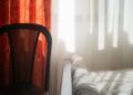 Sunlight streaming through curtains illuminating a bed and chair in a cozy bedroom setting.