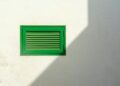 Green vent on a white wall with sharp sunlight casting a shadow, creating a minimalist urban scene.