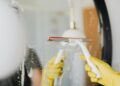 Anonymous person in yellow rubber gloves wiping water by squeegee from mirror in bathroom