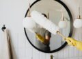 Crop anonymous female housewife in rubber gloves dusting modern lamp with soft brush against mirror while cleaning stylish bathroom