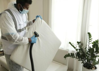 Man in protective gear vacuuming a mattress in a modern home interior.