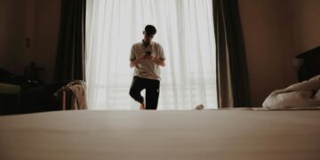 Young man stands by window in modern bedroom, using a smartphone.