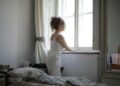 Back view of curly hair female wearing pajamas standing on bed and looking out of window after awakening in bedroom