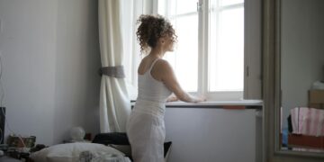 Back view of curly hair female wearing pajamas standing on bed and looking out of window after awakening in bedroom