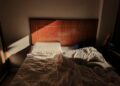 A sunlit, unmade bed with a wooden headboard creates a cozy and warm ambience.