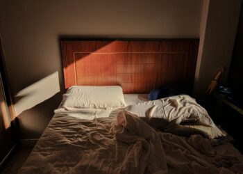 A sunlit, unmade bed with a wooden headboard creates a cozy and warm ambience.
