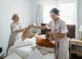 Two female housekeepers in uniform efficiently prepare a bed in a bright, well-lit room.