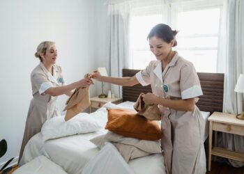 Two female housekeepers in uniform efficiently prepare a bed in a bright, well-lit room.