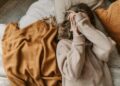 Woman in knitted sweater relaxing on bed with cozy blankets, covering face for warmth.