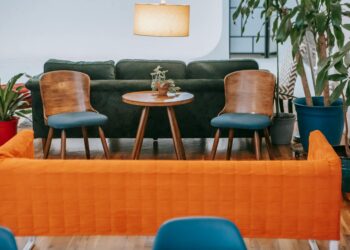 Comfortable sofa with wooden chairs near round table in stylish apartment with green potted plants