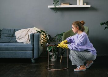 Side view of young female in latex gloves squatting down and wiping table in stylish living room in daytime