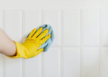 Crop anonymous housekeeper in yellow gloves cleaning white tiled wall using rag in bathroom