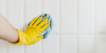 Crop anonymous housekeeper in yellow gloves cleaning white tiled wall using rag in bathroom