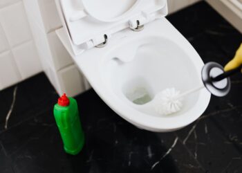 From above of crop anonymous person in rubber gloves using liquid toilet sanitizer and brush to clean toilet bowl in bathroom
