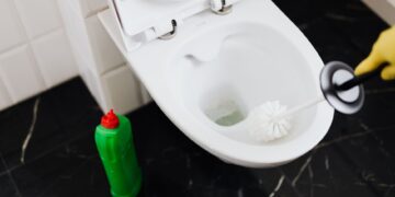 From above of crop anonymous person in rubber gloves using liquid toilet sanitizer and brush to clean toilet bowl in bathroom