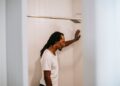 Side view of sad young informal black guy with Afro braids in white t shirt standing in shower booth under water stream and leaning on wall