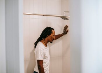 Side view of sad young informal black guy with Afro braids in white t shirt standing in shower booth under water stream and leaning on wall