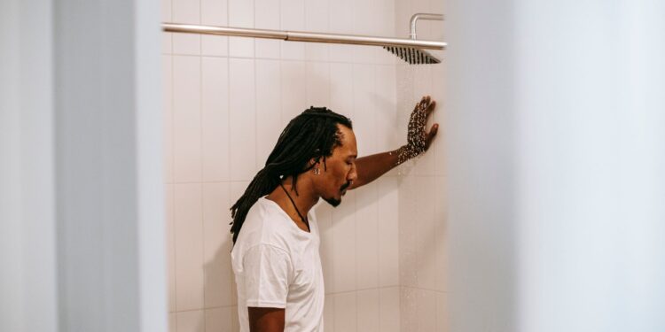 Side view of sad young informal black guy with Afro braids in white t shirt standing in shower booth under water stream and leaning on wall
