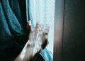 A hand gently opens a curtain, revealing sunlight streaming through a window indoors.