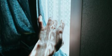 A hand gently opens a curtain, revealing sunlight streaming through a window indoors.