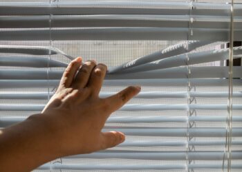 A hand gently opens window blinds, letting sunlight stream indoors.
